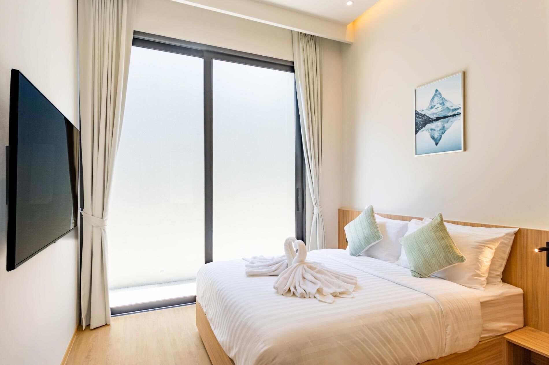 A hotel room with a bed, a TV, a large window with curtains, and a painting on the wall.