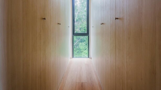 Un couloir étroit, doté de planchers et de portes en bois, qui mène à une fenêtre offrant une vue sur les arbres.