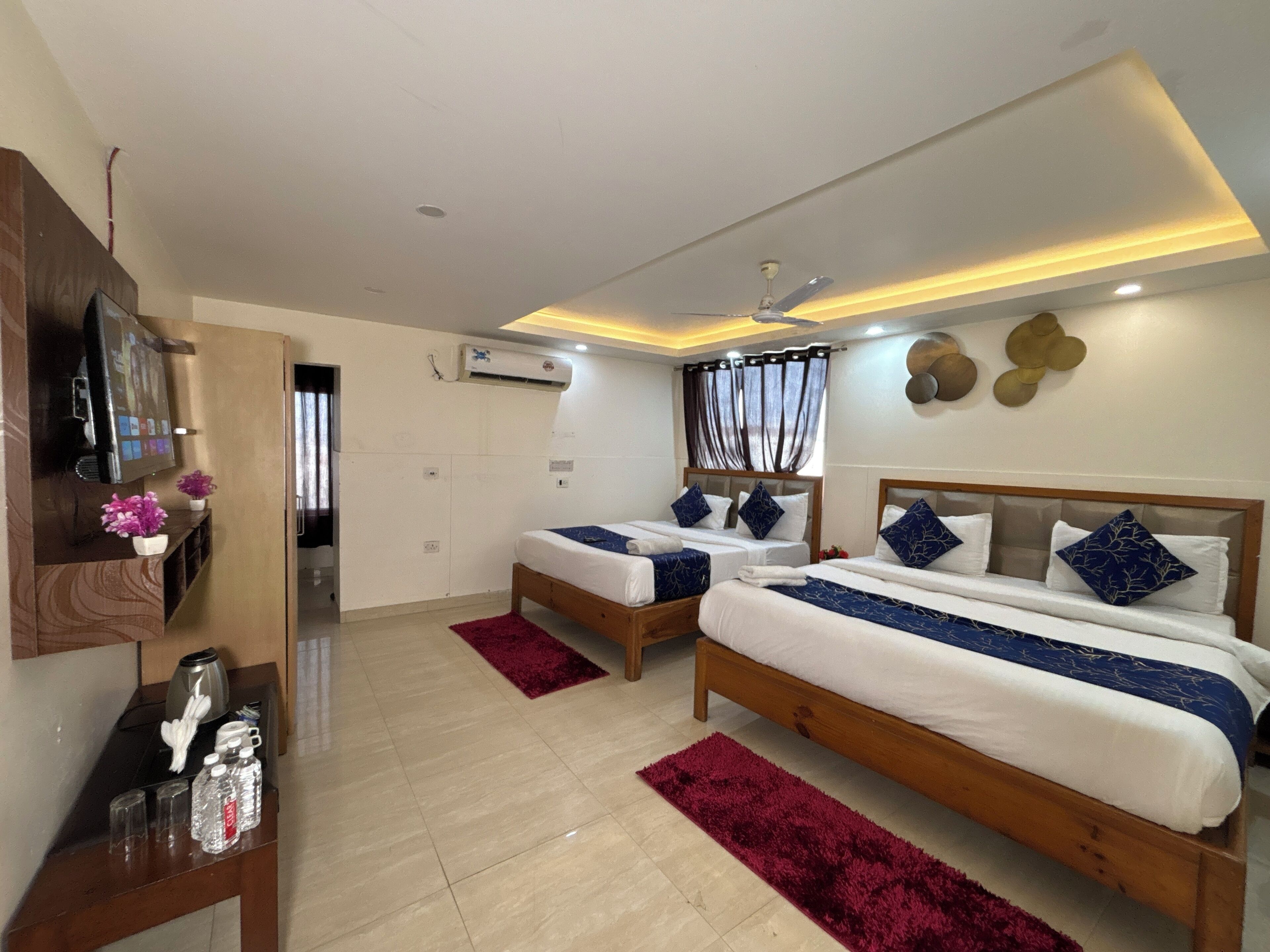 A hotel room with two beds, a TV, an air conditioner, and a wooden desk with a water bottle and glasses.