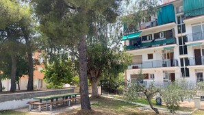 A multi-story residential building with balconies, a green bench, and a large tree in the foreground. - Ayres, Simple Apartment (El Vendrell)