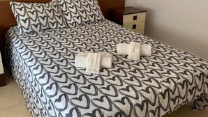 A bed with a heart-patterned bedspread, a wooden headboard, a nightstand with a drawer, and a folded towel on the bed. - Ayres, Simple Apartment (El Vendrell)