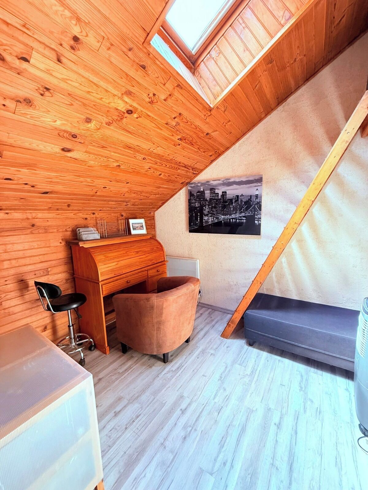 A loft bedroom with wooden slats on the ceiling, a cityscape picture on the wall, a desk with a chair, and a bed.