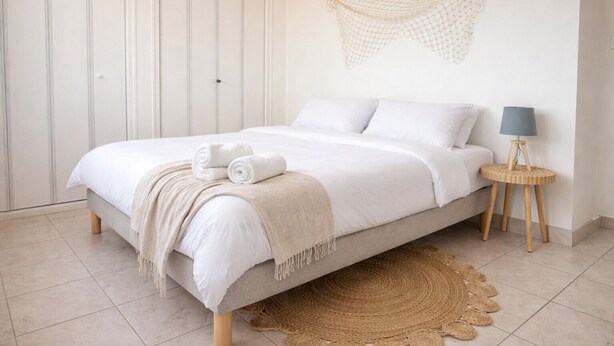 Een bed met een beige deken en opgerolde handdoeken, een rond houten nachtkastje met een lamp, en een geweven vloerkleed.