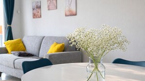 Een eetgedeelte met een ronde tafel, teal stoelen, een vaas met witte bloemen, een grijze bank met gele kussens en kunst aan de muur.