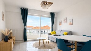 Een moderne woonkamer met een bank, een eettafel met stoelen, een ronde salontafel, een houten bijzettafeltje en een uitzicht op gebouwen door grote ramen.