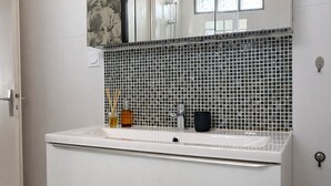 Een badkamer met een wit wastafelmeubel, een spiegel, een betegelde spatwand en een raam.