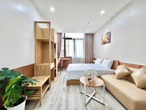 Une chambre d’hôtel avec un lit, un canapé, un bureau, une chaise, une plante en pot et une fenêtre avec des rideaux.
