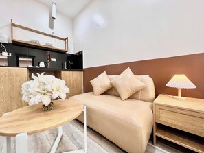 A modern room with a sofa, a small table with a vase of flowers, a lamp, and a kitchen area in the background. - Aurora Homestay 16 Nguyen Nhu Do (Dong Da)