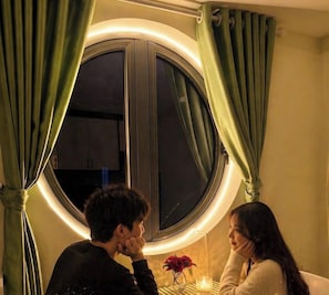 Una habitación con un ventanal grande, cortinas, una mesa con una vela y un florero con flores.