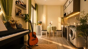A compact living space with a piano, a guitar, a dining table with chairs, a washing machine, and a small kitchen area. - La Yen Homes (Ho Chi Minh City)