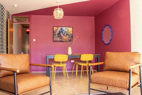 A room with a dining table, two chairs, a painting, and a vase with flowers.