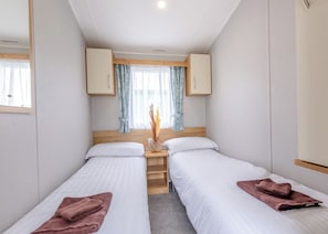 A bedroom with two single beds, a wooden headboard, a nightstand with a vase, and a window with curtains. - Bronze 2 Bedroom Caravan, Sleeps 4 (Great Yarmouth)