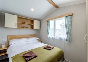 Ein Schlafzimmer mit einem Bett, einem Nachttisch, einem Holzregal über dem Bett, einem Fenster mit Vorhängen und einer Steckdose.