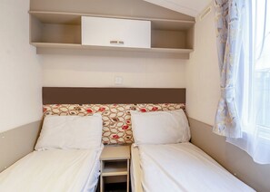 Een kleine, eenvoudige kamer met twee bedden, een plank erboven en een raam met gordijnen.
