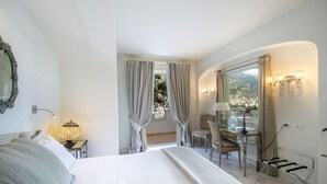 A hotel room with a bed, a desk, a chair, a lamp, a mirror, and a view of a balcony. - Villa Isea Positano (Positano)
