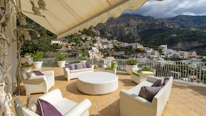 A terrace with white furniture, potted plants, and a view of a mountainous town. - Villa Isea Positano (Positano)