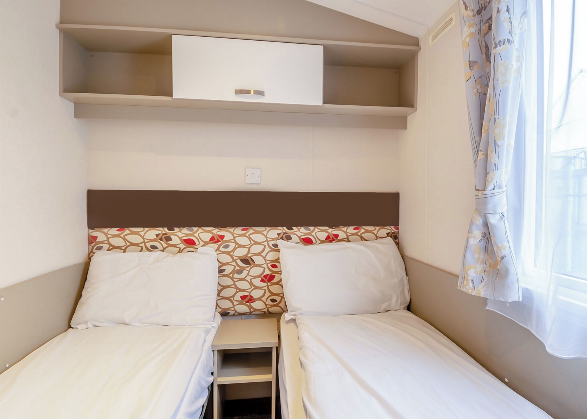 A small, compact room with two single beds, a small shelf above the beds, and a window with curtains.