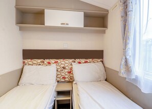 A small, compact room with two single beds, a small shelf above the beds, and a window with curtains. - Gold Caravan with parking & decking P (Great Yarmouth)