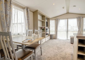 A modern room with a dining table, chairs, a bookshelf, and a TV. - Gold Caravan with parking & decking P (Great Yarmouth)