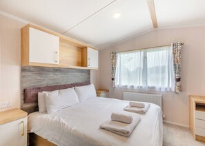 Un dormitorio con cama, cabecero de madera, ropa de cama blanca, una ventana con cortinas y una mesita de noche.