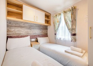A bedroom with two beds, wooden headboard, white curtains, and a small bedside table. - Gold Premium Caravan with ensuite & decking 6 P (Great Yarmouth)
