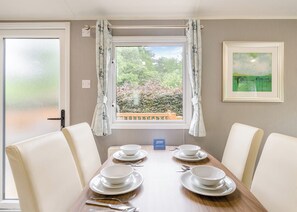 A dining area with a table set for four, a window with curtains, a framed painting, and a sign indicating a restaurant. - Gold Premium Caravan with ensuite & decking (Great Yarmouth)