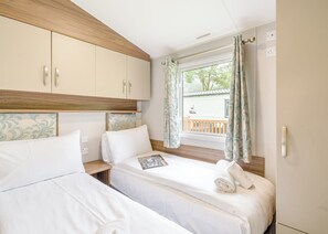 A bedroom with two beds, wooden headboard, a window with curtains, and a view of a balcony. - Gold Premium Caravan with ensuite & decking (Great Yarmouth)