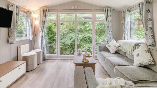 Ein gemütliches Zimmer mit einer Couch, einem kleinen Tisch, einem Fernseher und großen Fenstern mit Vorhängen.