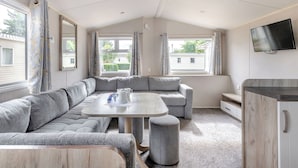 A modern RV interior with a sectional sofa, a round table, and a TV mounted on the wall. - Silver Caravan with decking 6 (Great Yarmouth)