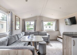 A modern RV interior with a sectional sofa, a round table, and a TV mounted on the wall. - Silver Caravan with decking 6 (Great Yarmouth)