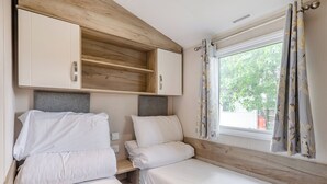 A compact bedroom with two beds, wooden headboards, a window with curtains, and built-in cabinets. - Silver Caravan with decking 6 (Great Yarmouth)