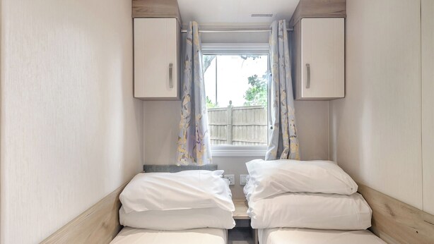 A small, single-room accommodation with two beds, a window with curtains, and a wooden headboard. - Silver Caravan with ensuite P (Great Yarmouth)