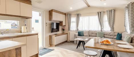 A modern kitchen with a built-in oven, a dining area with a wooden table and bar stools, a sofa, and a TV mounted on the wall.