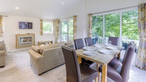 Een ruime woonkamer met een open haard, een bank, een eettafel gedekt voor vier personen en grote ramen met uitzicht op groen.