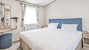 A bedroom with a bed, a nightstand, a mirror, a window with curtains, and a radiator. - Gold Accessible with parking P (Great Yarmouth)