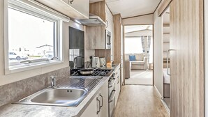 A compact kitchen with a sink, stove, and microwave, a window with a view of parked cars, and a door leading to a living area with a sofa. - Gold Accessible with parking P (Great Yarmouth)