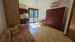 Ein Wohnzimmer mit einer Couch, einem Esstisch mit Stühlen, einer Küchenzeile mit Schränken und einer Glastür zum Außenbereich.