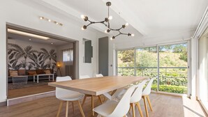 Et moderne spiseområde med et træbord og hvide stole, en lysekrone samt udsigt til grøn vegetation gennem store vinduer.