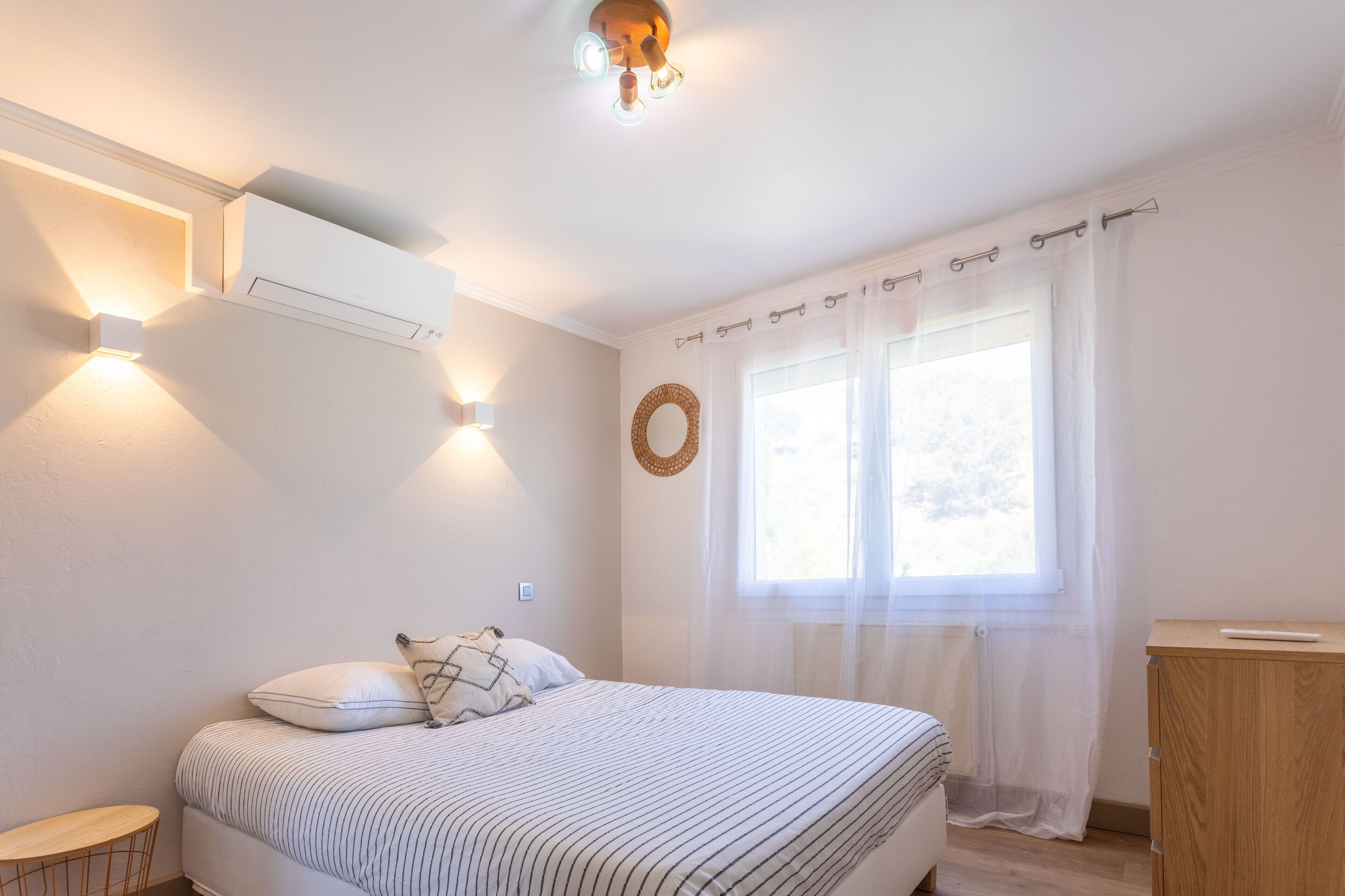 Quarto com cama, mesa de cabeceira, ar condicionado fixado na parede e janela com cortinas leves.