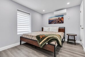 A bedroom with a bed, a window with blinds, an air conditioning unit, a small round table, and a painting on the wall. - Beach Block, 2BR, Free Parking in Seaside Heights (Seaside Heights)