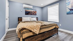 A bedroom with a bed, a painting, a window with blinds, and an air conditioning unit. - Charming Seaside Escape | 2BR, Parking, Beach (Seaside Heights)