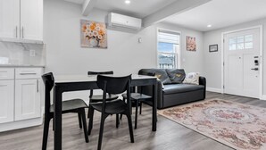 A modern living room with a dining table, chairs, a sofa, and a large window. - Charming Seaside Escape | 2BR, Parking, Beach (Seaside Heights)