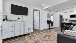 A modern living room with a flat-screen TV, white cabinetry, a dining area with black chairs, and a kitchen with white cabinets. - Charming Seaside Escape | 2BR, Parking, Beach (Seaside Heights)