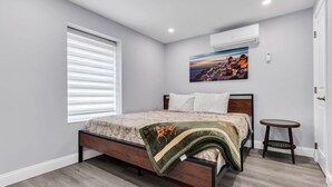 Ein Schlafzimmer mit Bett, einem Fenster mit Jalousien, einer Klimaanlage, einem kleinen runden Tisch und einem Gemälde an der Wand.