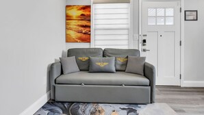 A grey sofa with matching cushions, a painting of a sunset, a white door, and a small framed item on the wall. - 1BR, Free Parking, Steps to the Boardwalk (Seaside Heights)