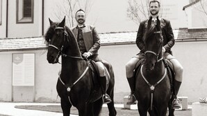 Dos personas a caballo frente a un edificio con techo de tejas y ventanas arqueadas.