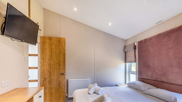 A bedroom with a bed, a wooden wardrobe, a television, a radiator, and a window with curtains. - Deluxe 2, Slps 4, parking, ensuite, decking, HT (Burgh Castle, Great Yarmouth)
