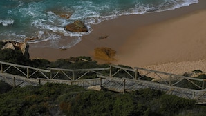 Vista costera con un camino de madera que conduce a una playa de arena y afloramientos rocosos.