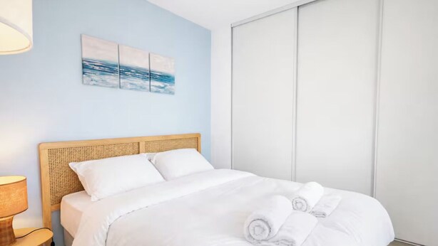 Un lit bien fait, avec du linge de lit blanc et des serviettes pliées, une tête de lit en bois, une table de chevet avec une lampe, et une œuvre d’art accrochée au mur au-dessus du lit.