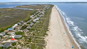 Vista aérea de uma área costeira com uma praia extensa, uma estrada paralela à costa e uma fileira de casas, com um edifício de telhado vermelho em destaque.
